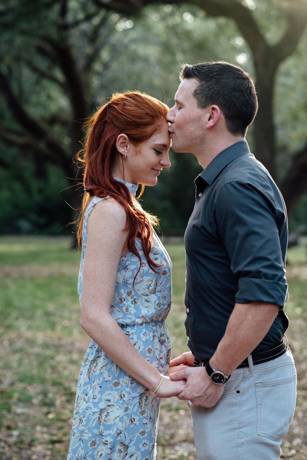 A man kisses a redheaded woman on the forehead while holding hands in a lush green park.