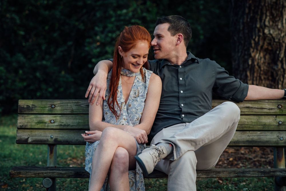 Happy couple sitting close on a park bench, smiling at each other in a lush green outdoor setting.