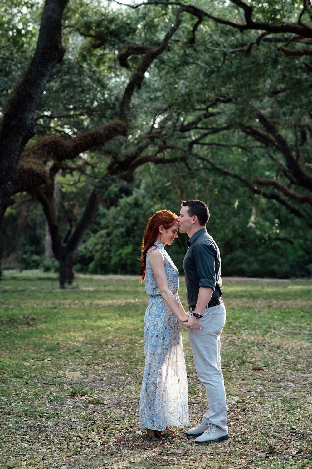 A couple shares a tender forehead kiss while holding hands in a scenic park with large oak trees.
