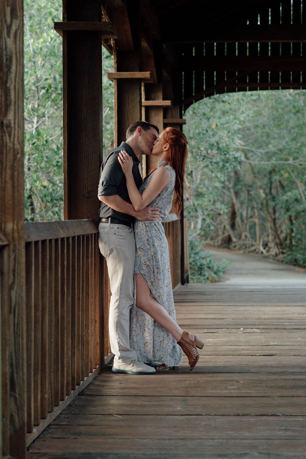 A couple shares a romantic kiss on a wooden bridge walkway surrounded by lush green trees.