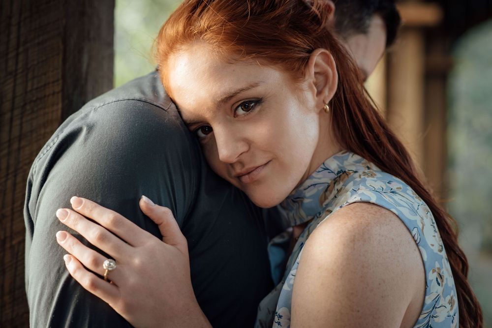 A red-haired woman with freckles hugs a man tenderly, smiling softly while wearing a floral dress and gold ring.