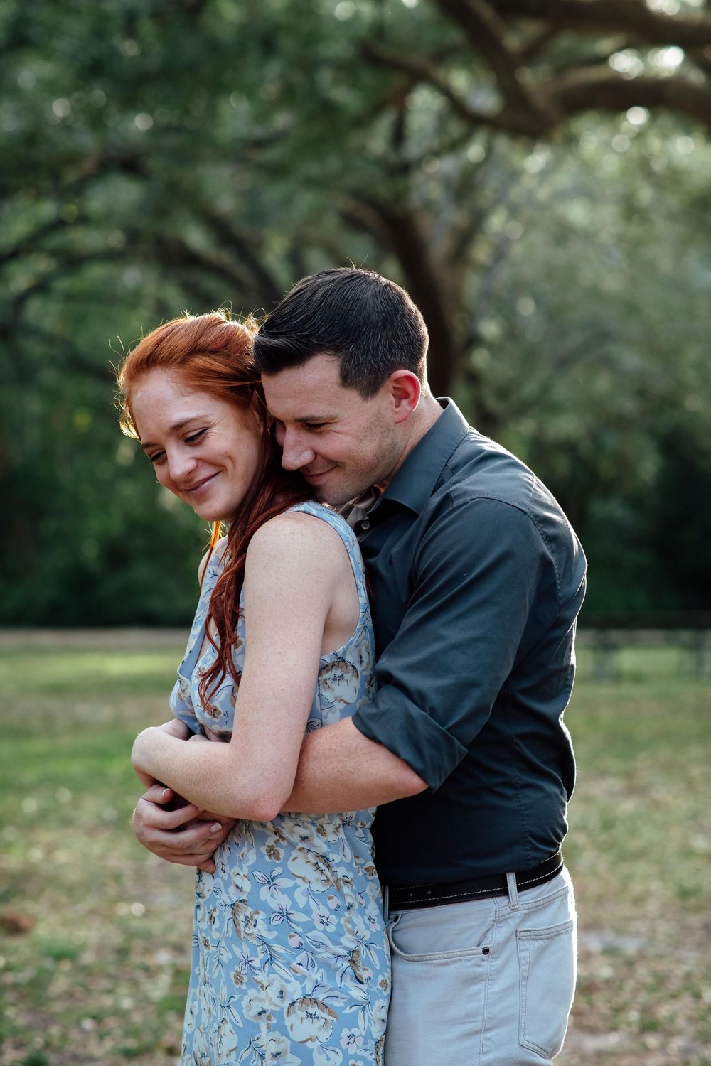 Couple embracing in a lush green park, woman in floral dress smiling as man hugs her from behind.