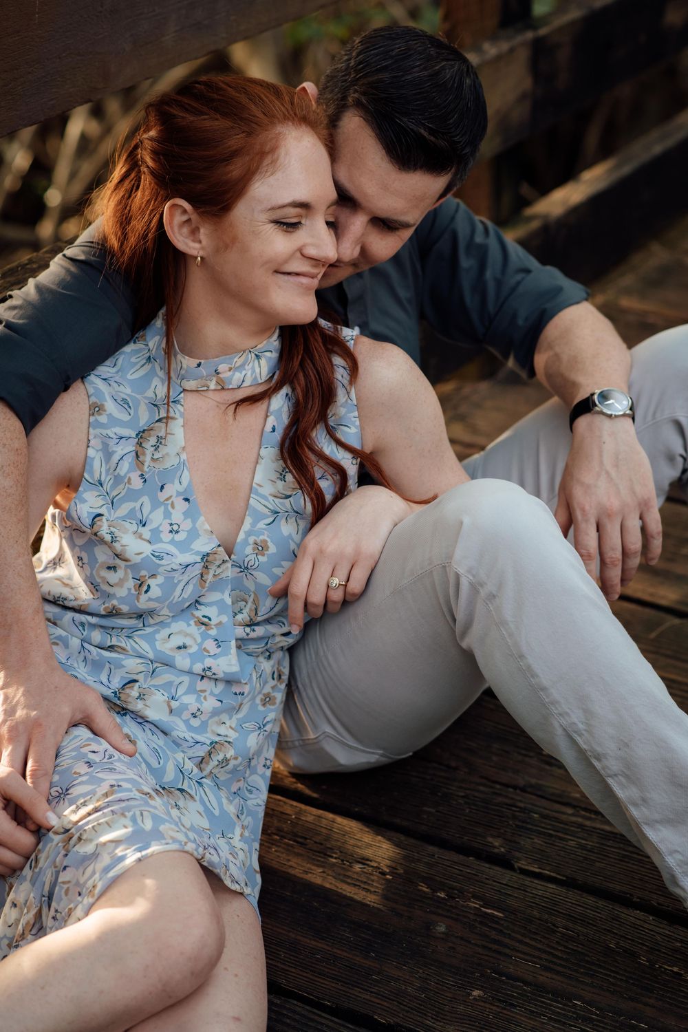 A smiling redhead woman in a blue floral dress sits closely with a man on a wooden deck.