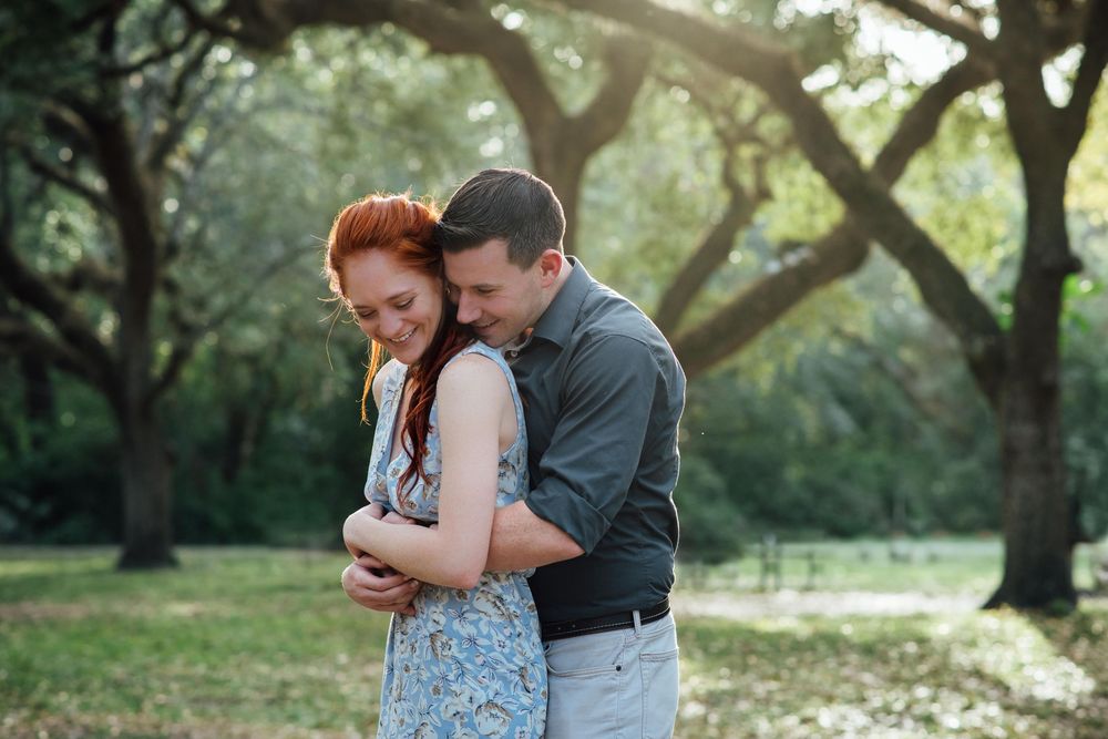 Couple embracing outdoors under lush green trees, woman with red hair smiling as man hugs her from behind.