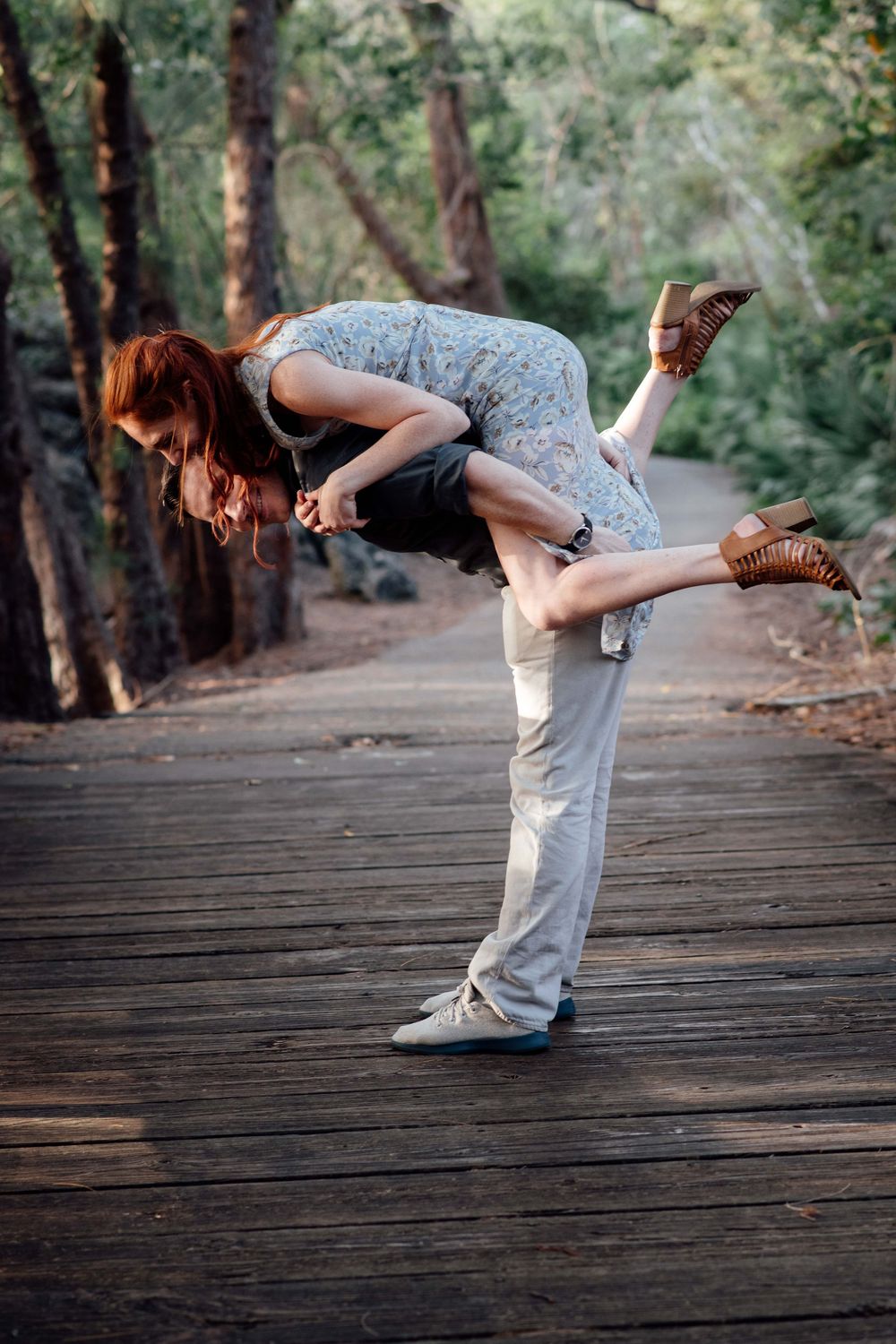 Man lifting woman in a romantic dip on a wooden path surrounded by trees.