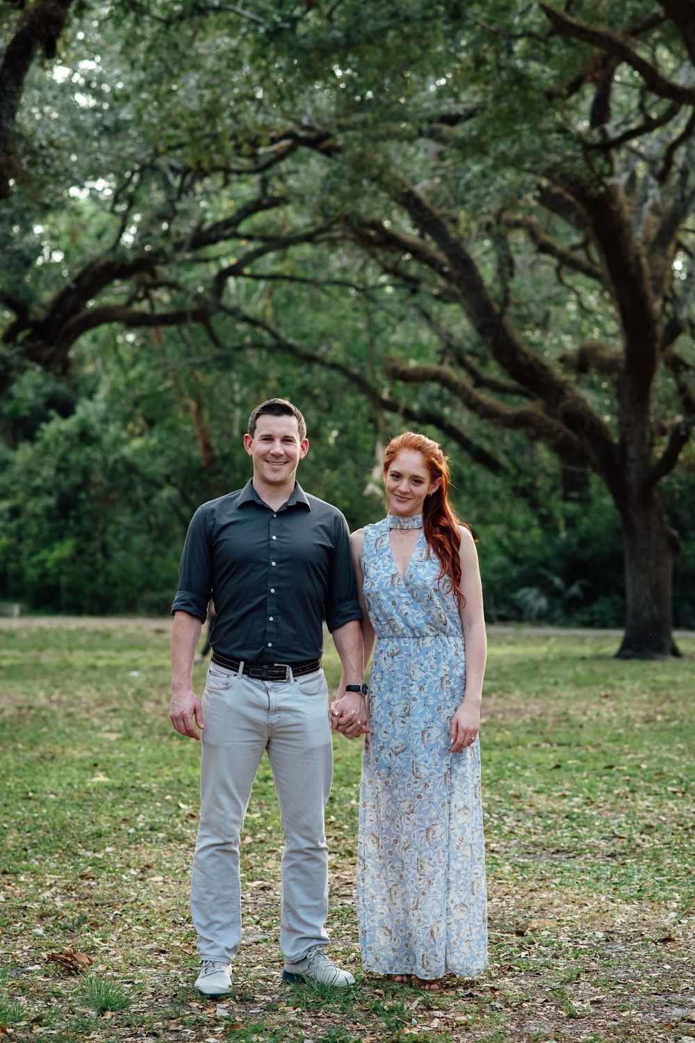 A couple holds hands smiling in a lush green park, man in dark shirt and khakis, woman in floral maxi dress.
