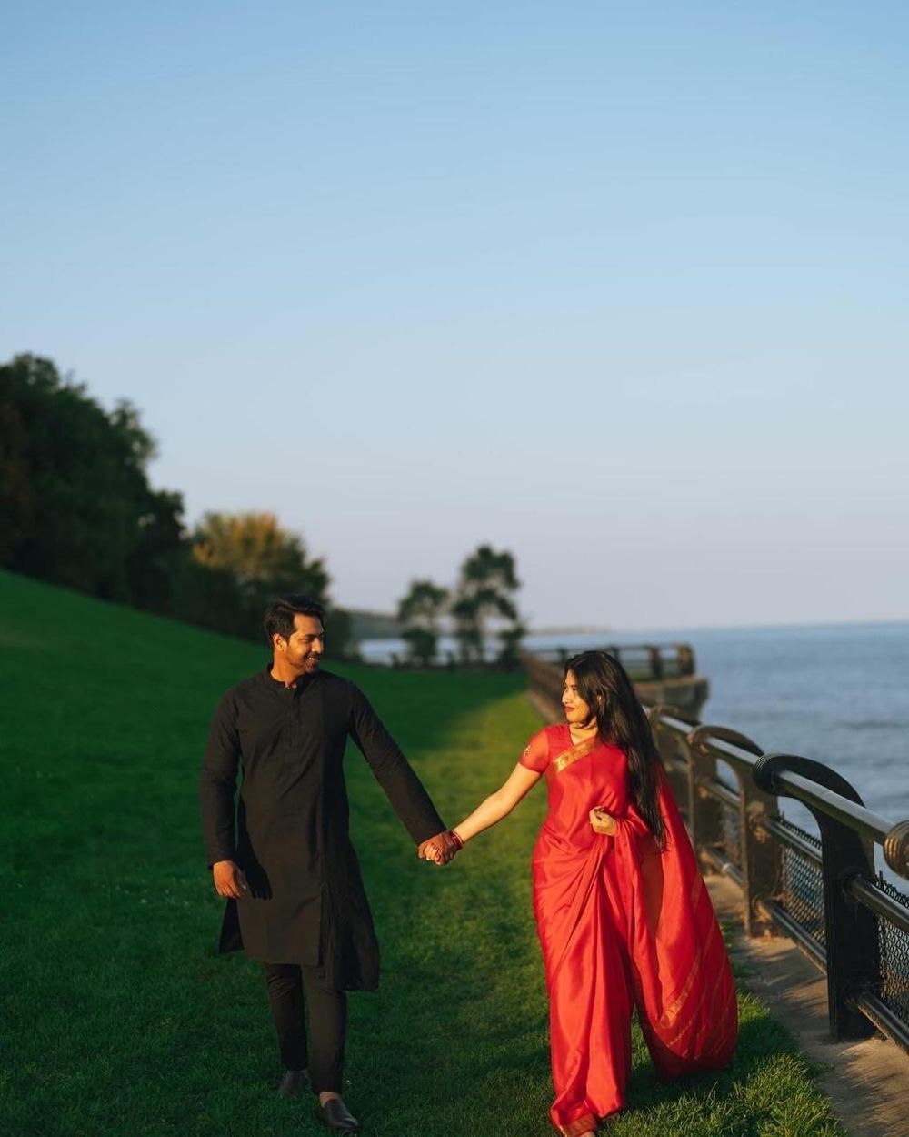 Couple in traditional Indian attire holding hands on green lawn by waterfront at sunset.