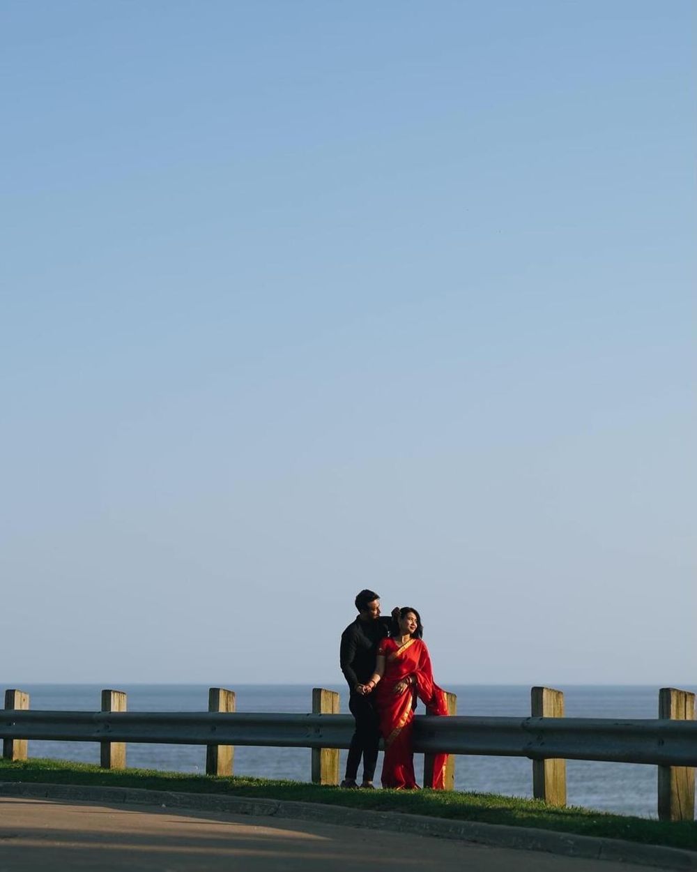 Couple in red and black traditional Indian attire posing by a coastal guardrail with ocean views.