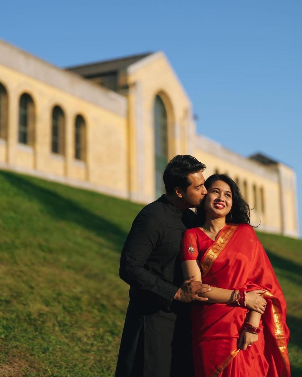Couple in red saree and black kurta sharing romantic moment on green hill with historic building backdrop.