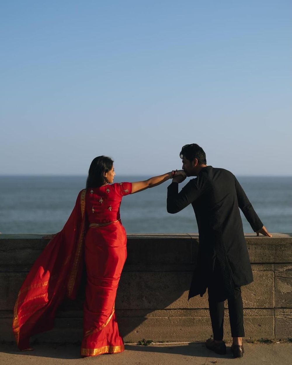 Couple in traditional Indian attire at seaside, man kissing woman's hand, red saree and black kurta.