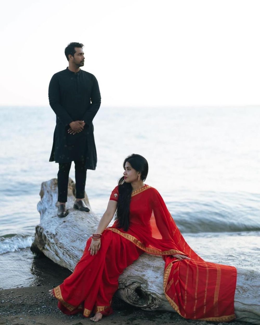 Indian couple in traditional attire, man in black kurta standing as woman in red saree sits on driftwood by the sea.