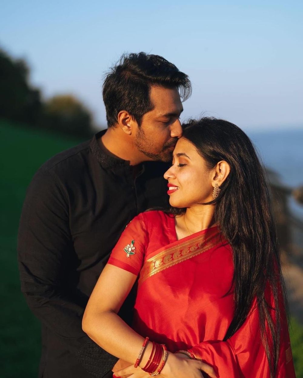 A couple in traditional South Asian attire share a tender moment outdoors, man in black kissing woman's forehead.