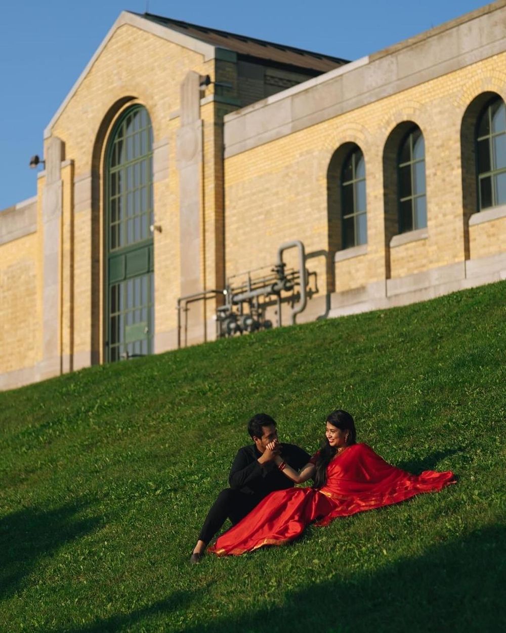 Couple in red and black attire sitting on a green hillside in front of a yellow brick building.