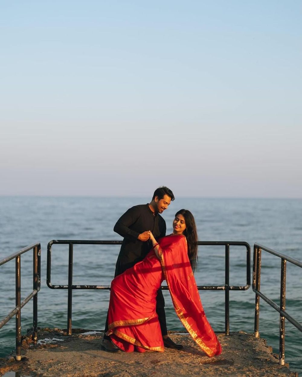 A couple in traditional Indian attire poses romantically on a seaside pier at sunset.
