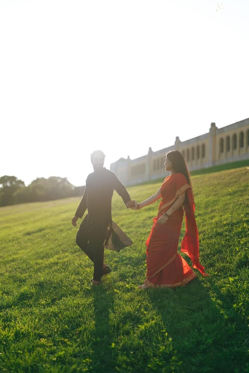Couple in traditional Indian attire holding hands walking on green grass at sunset near a historic building.