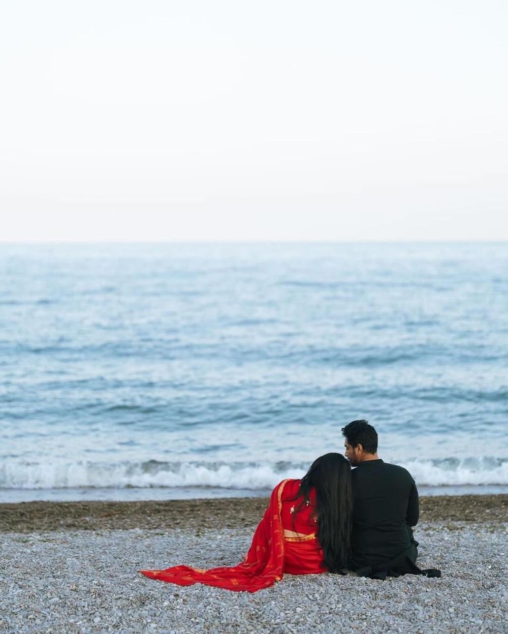 A couple in red saree and black outfit sits romantically on a pebble beach gazing at the ocean waves.