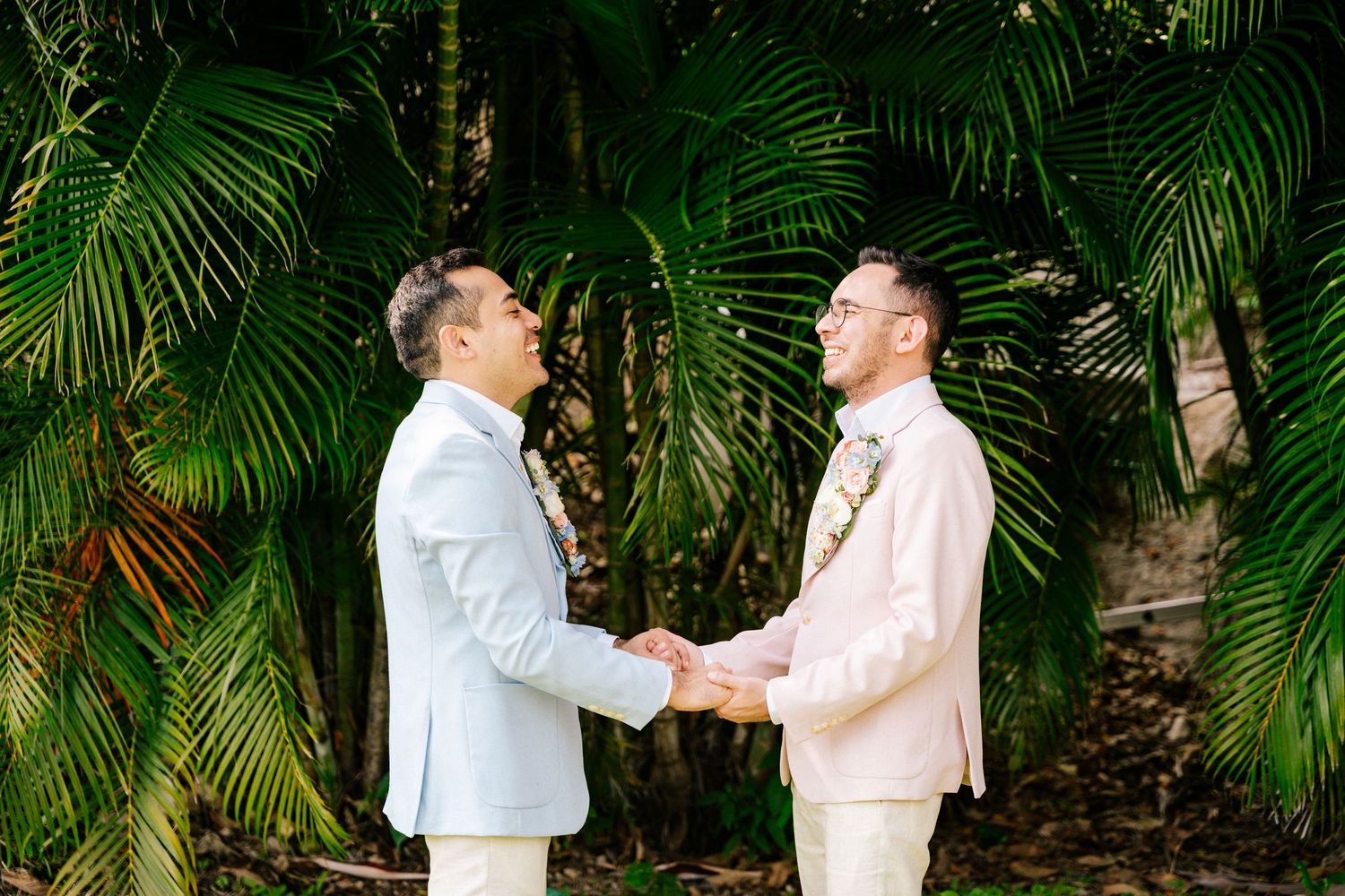 Two grooms in light suits hold hands during an outdoor wedding ceremony surrounded by tropical palm trees.