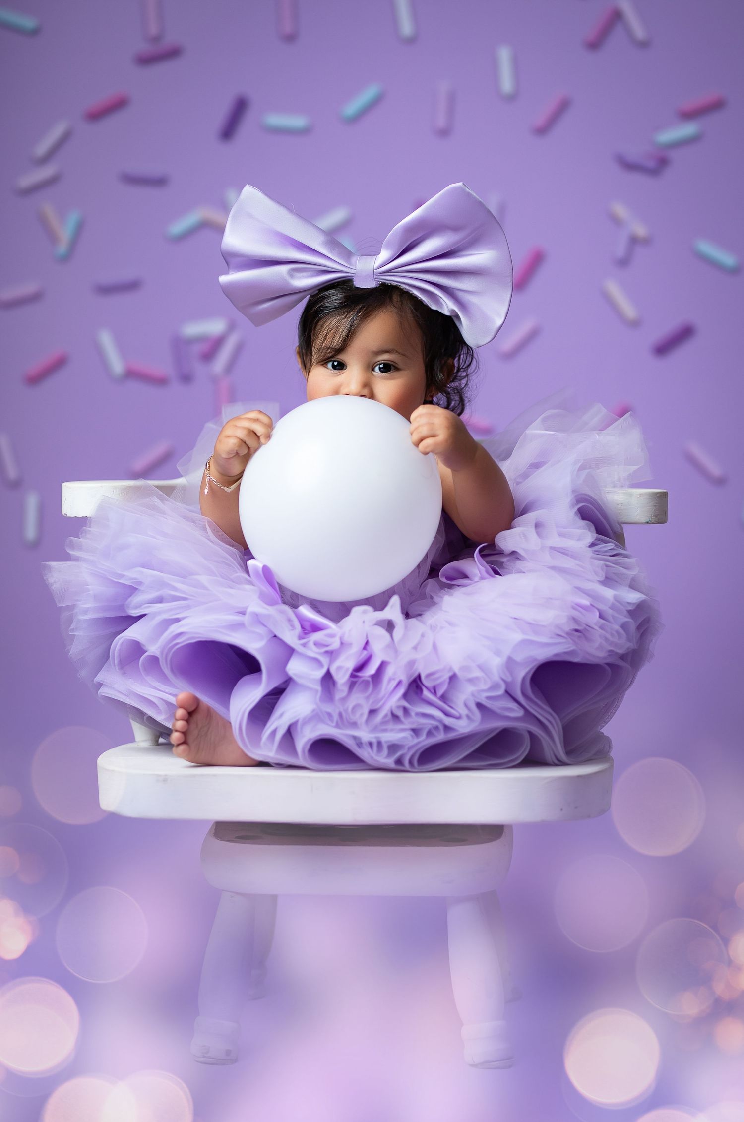 Baby girl in yellow tutu dress sits on white box before pink floral backdrop with large bow.