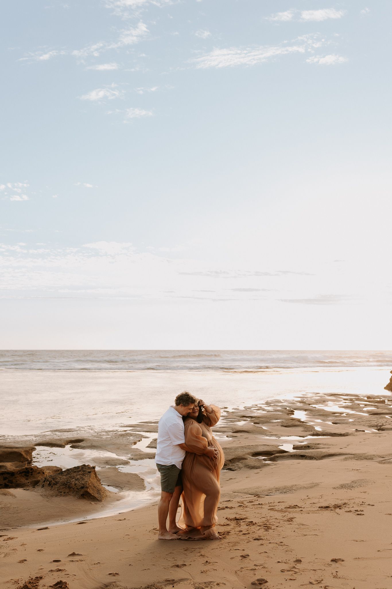 A couple embraces romantically on a sandy beach at sunset with calm ocean waves in the background.