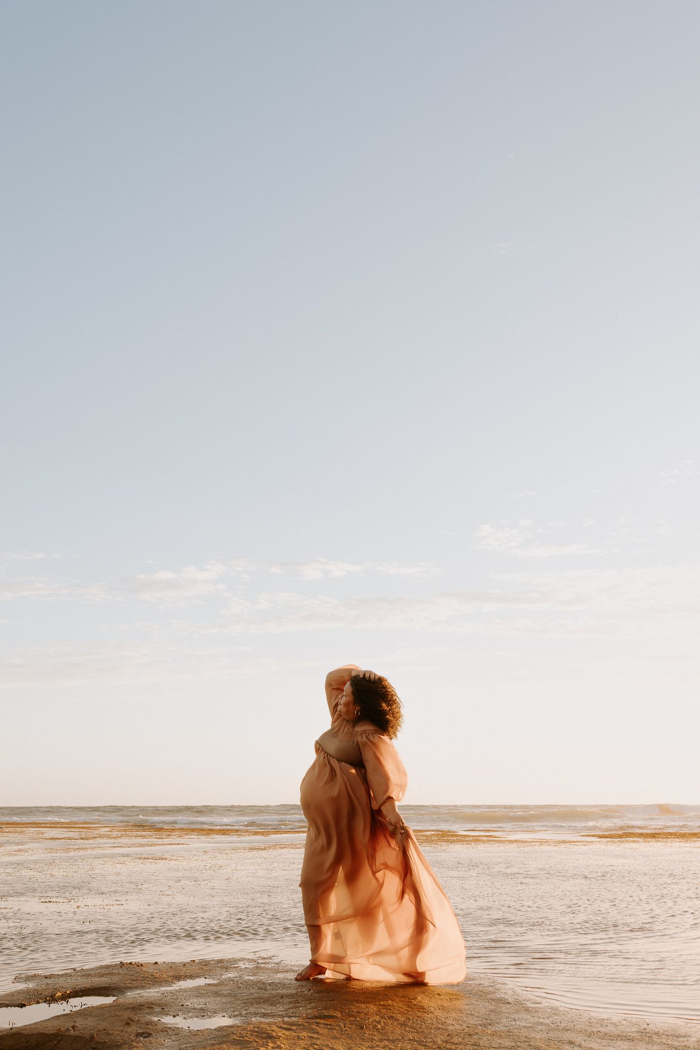 Pregnant woman in flowing peach dress standing on a serene beach at golden hour, cradling her baby bump.