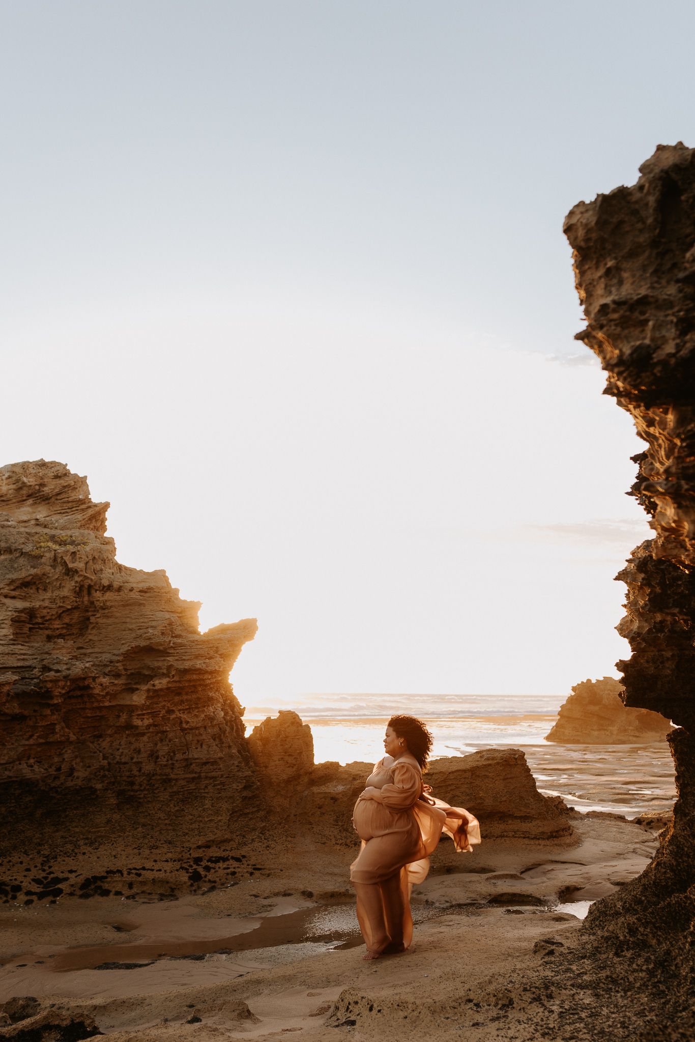 Pregnant woman standing between dramatic rocky cliffs on a sandy beach at golden hour, with ocean waves in the background.