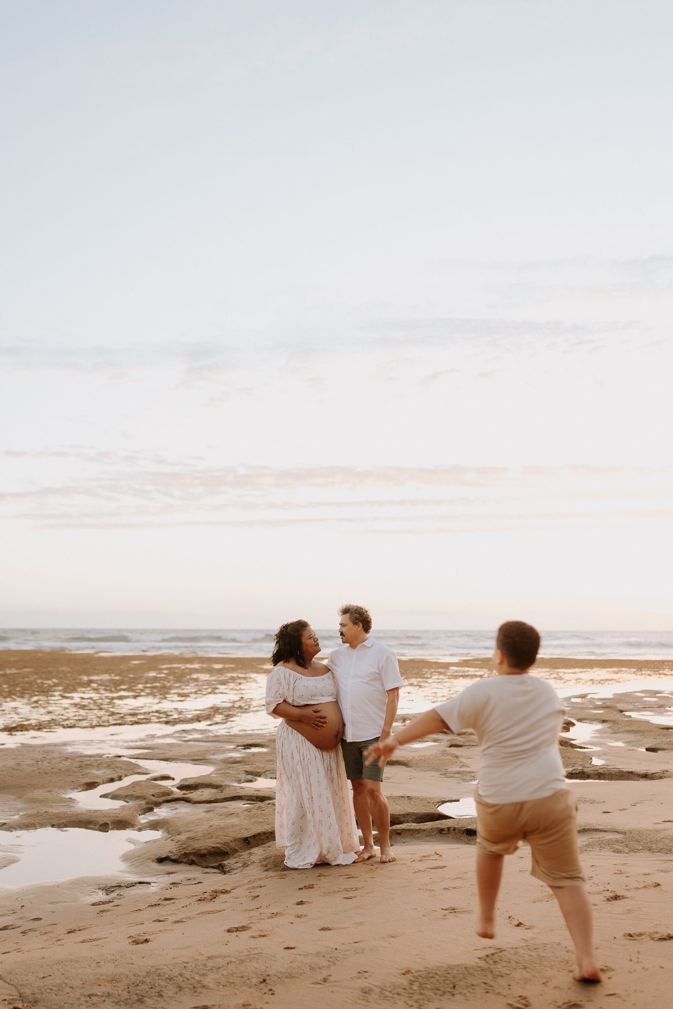 Photographer capturing a couple on a sandy beach at sunset, woman in white dress holding her baby bump.
