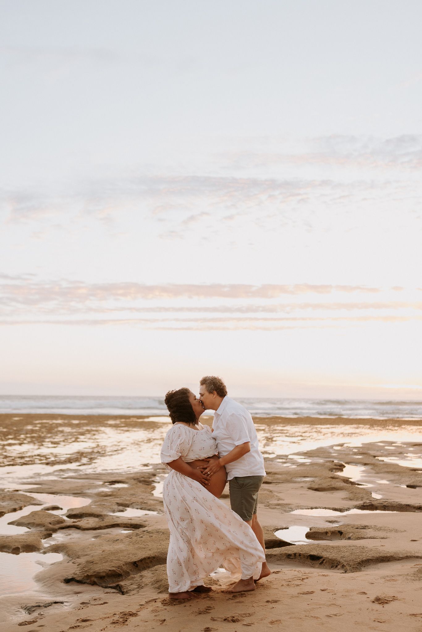 Couple sharing a romantic kiss on a sandy beach at sunset, woman in flowing white dress, waves in the background.
