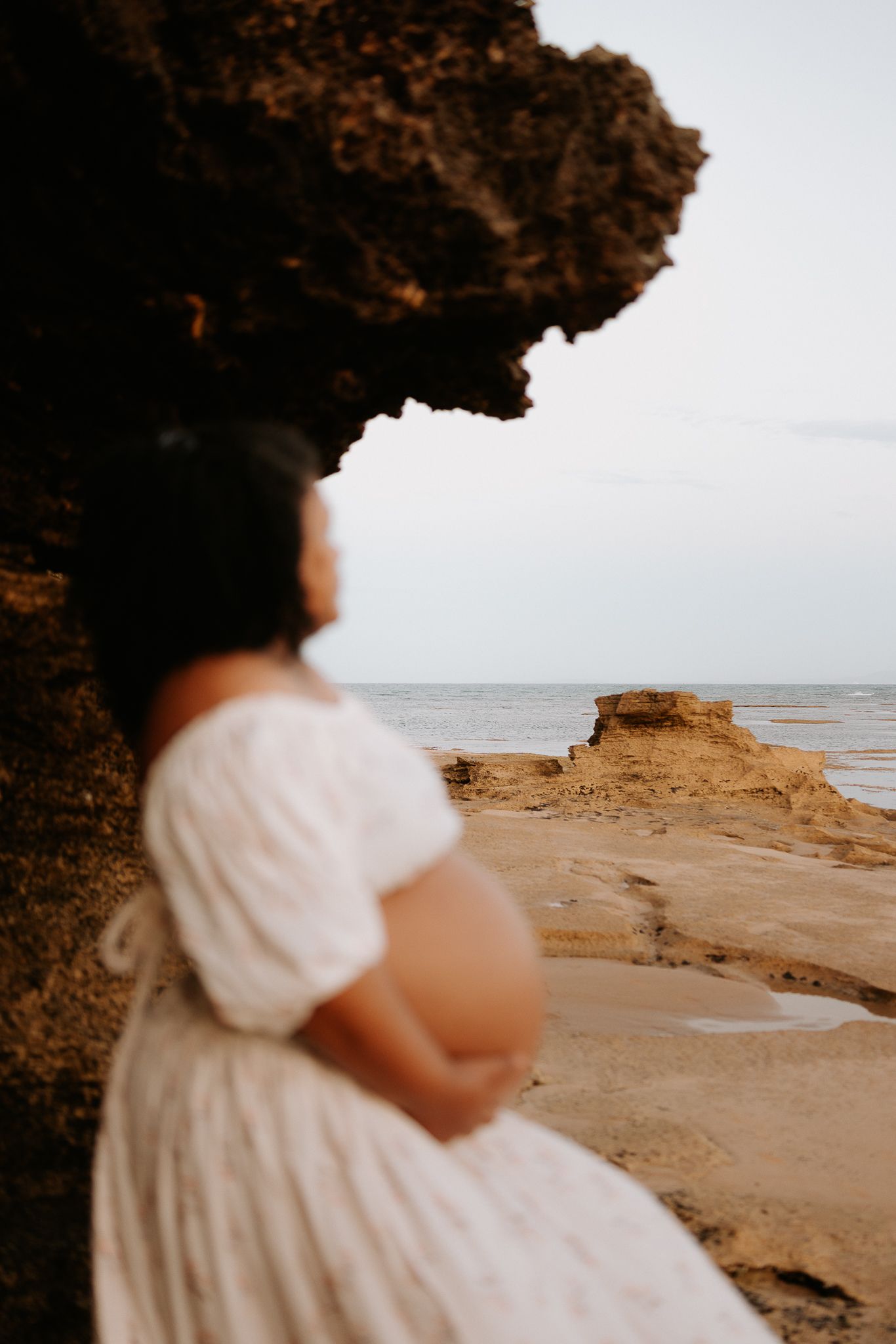 Pregnant woman in white dress standing by rocky coastal cliffs, gazing at the ocean during golden hour.