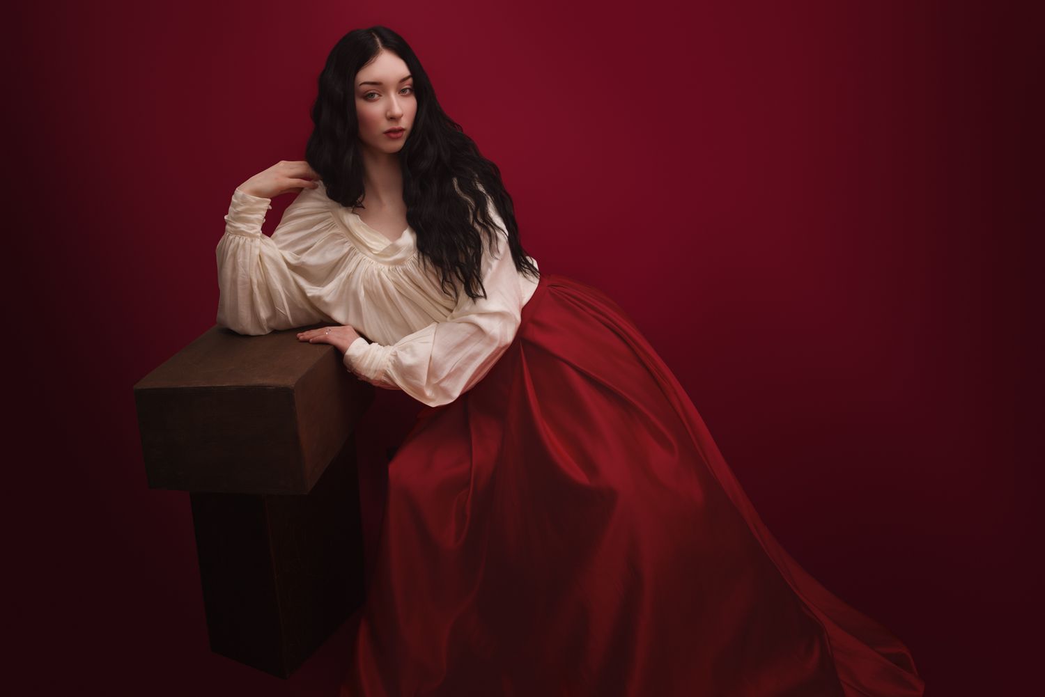Woman in red flowing skirt and white blouse poses elegantly against a deep red background in a classical portrait style.