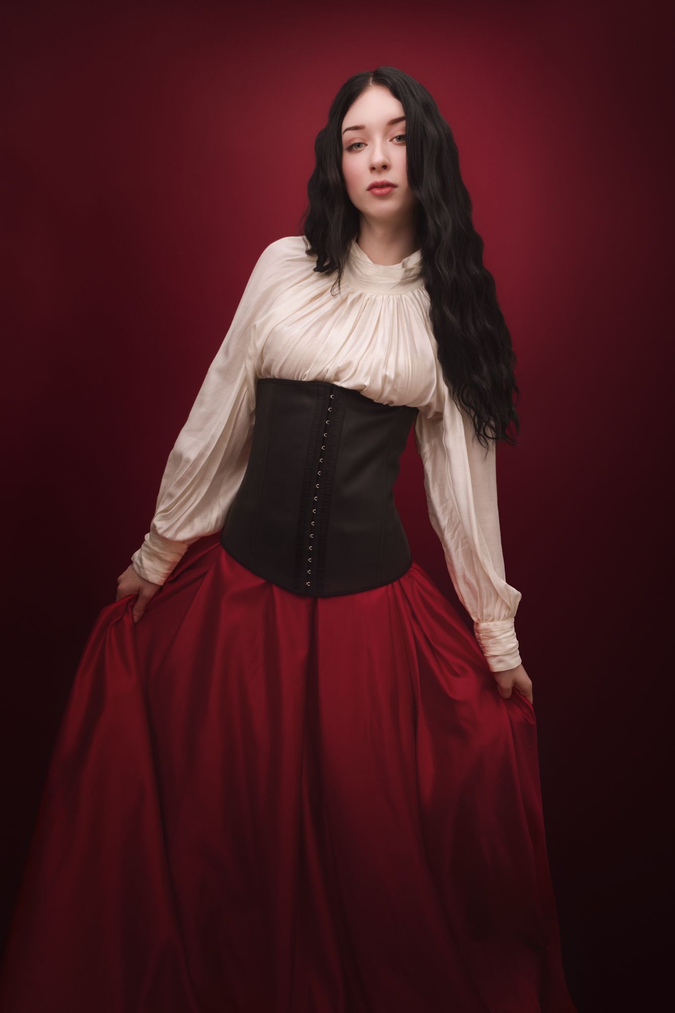 Woman in Victorian-style white blouse, black corset, and red skirt posing against a dark red background.