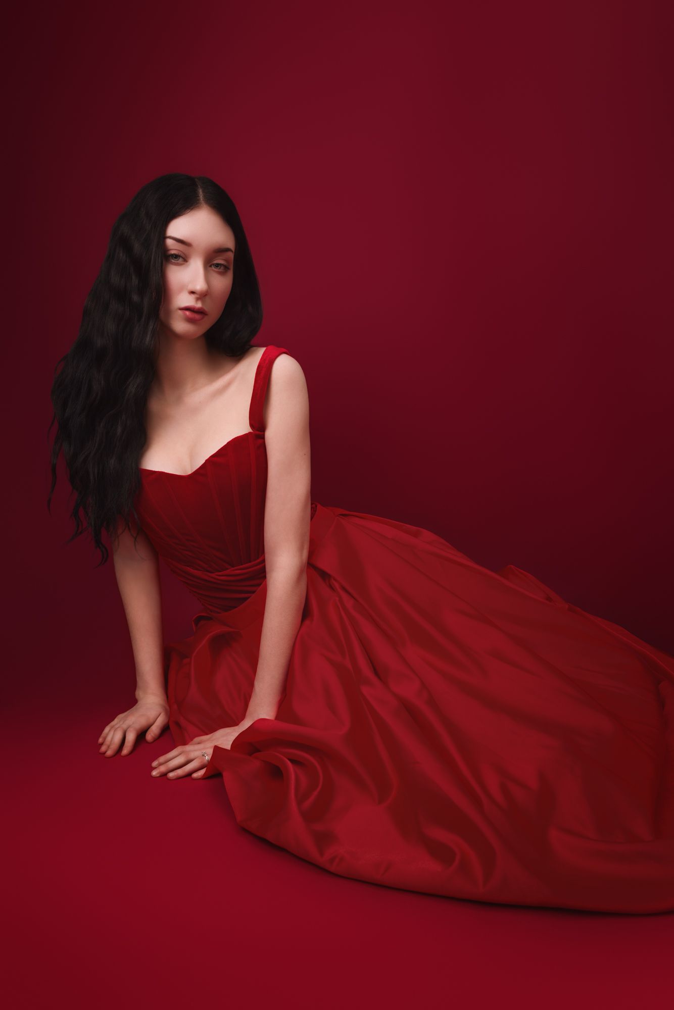 A woman in a flowing red gown sits gracefully on the floor against a deep red background with long dark wavy hair.