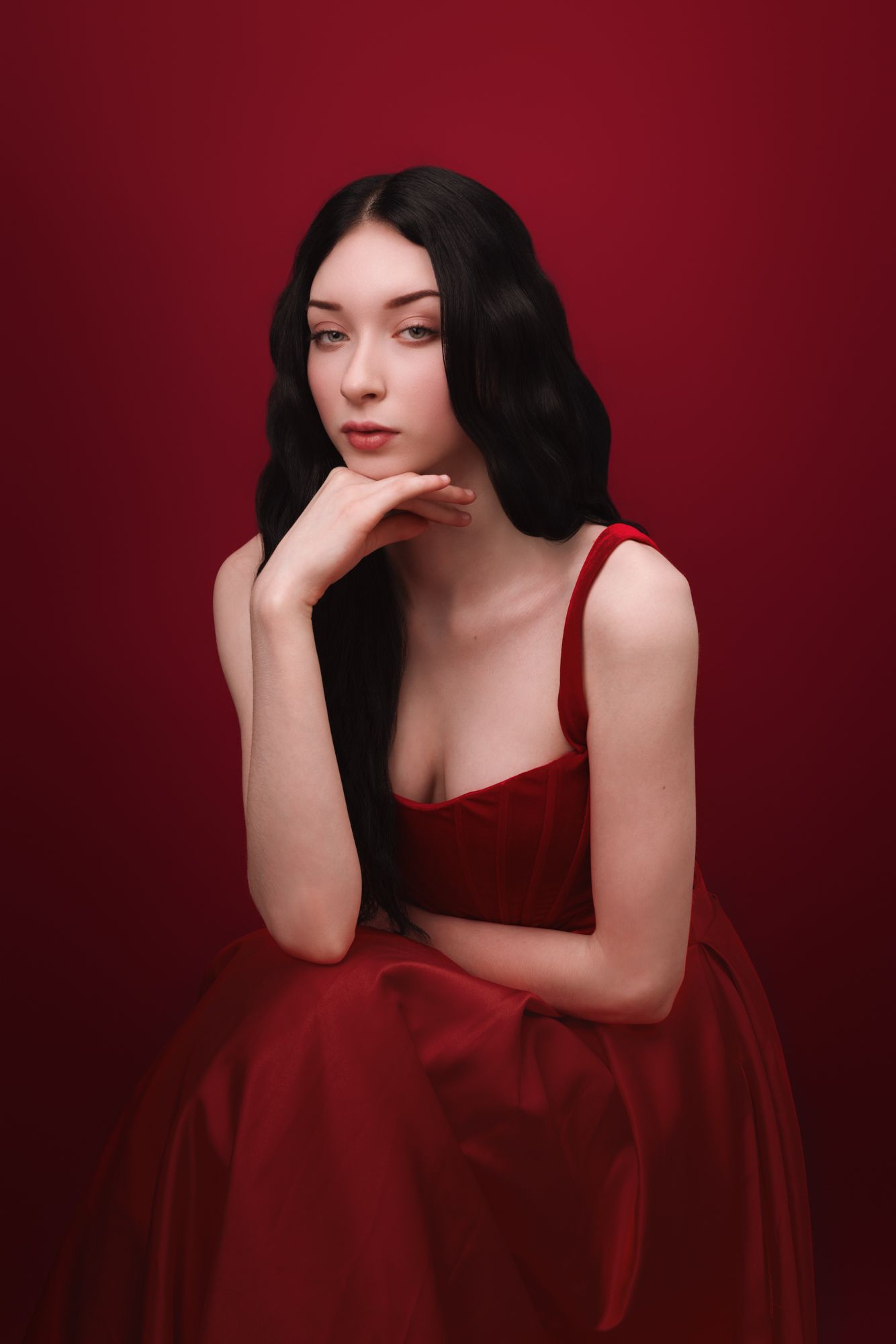 Young woman with black wavy hair in a red velvet gown poses gracefully against a deep red background.