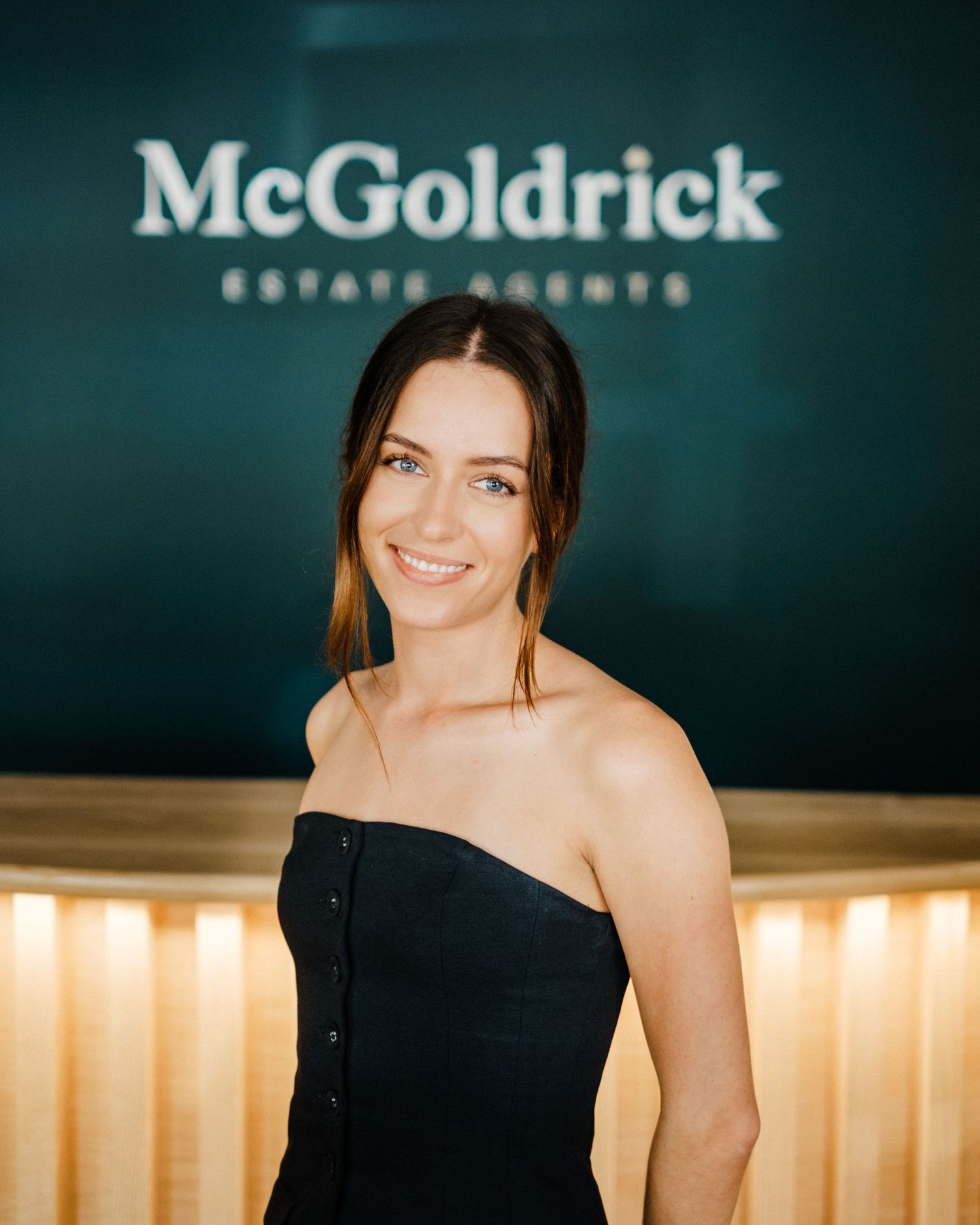Woman in black strapless dress smiling in front of McGoldrick Estate Agents office reception.
