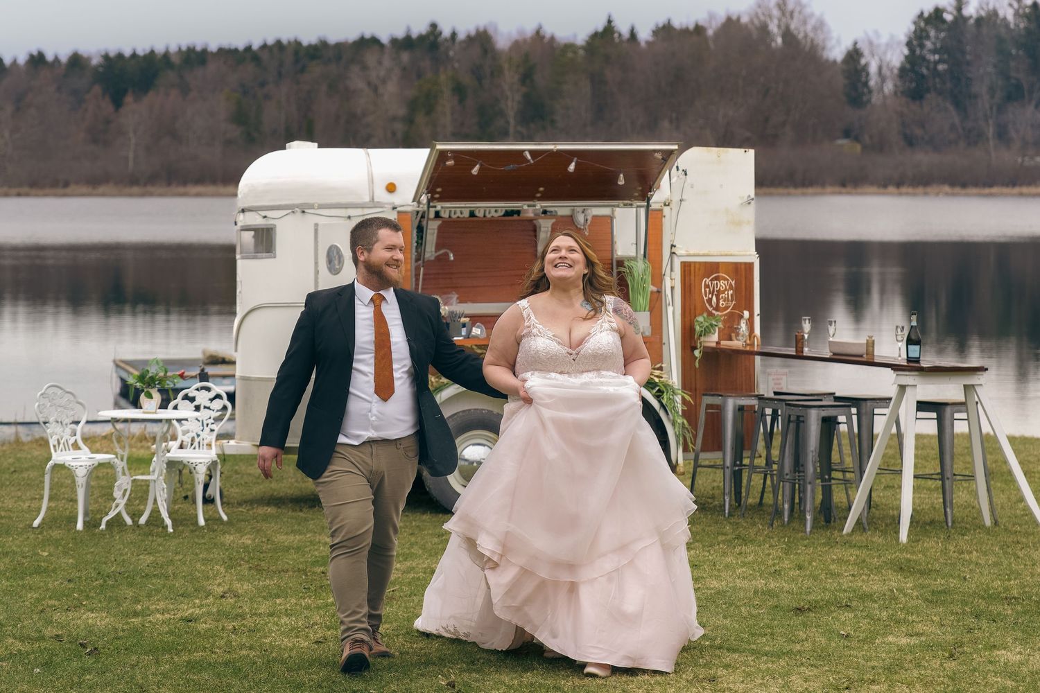 Bride and groom walking happily outdoors near a lakeside vintage food truck at their rustic wedding celebration.
