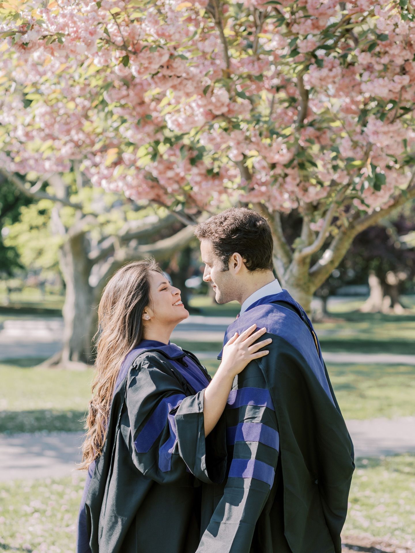 Two graduates in academic robes share a tender moment under a blooming pink cherry blossom tree in a sunny park.