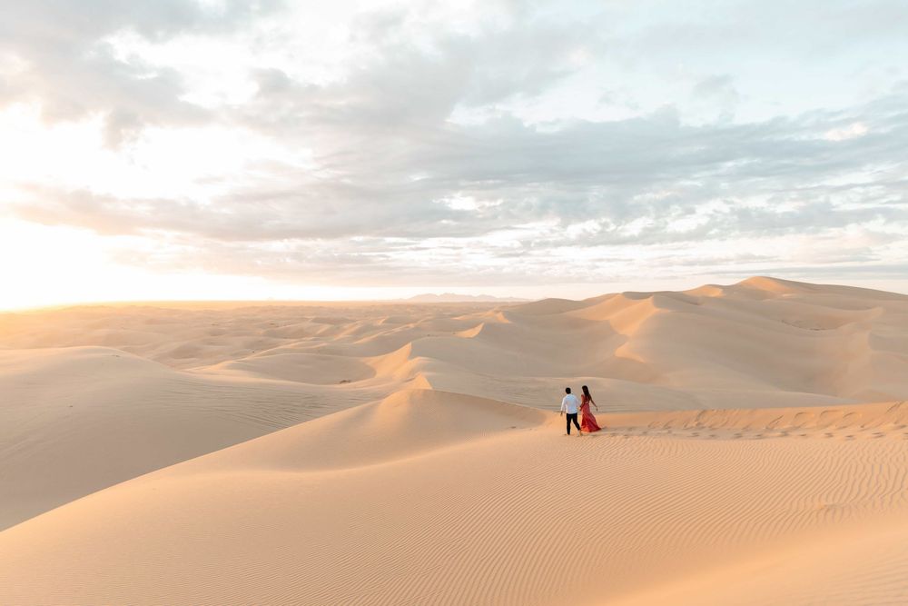 Couple standing on vast golden sand dunes at sunset in a sweeping desert landscape.
