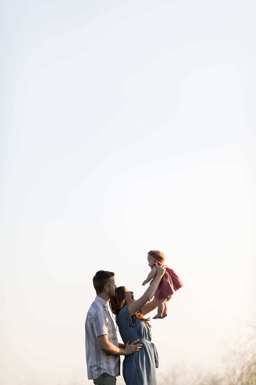 Parents lifting baby girl in pink dress against clear sky in joyful family portrait outdoors.