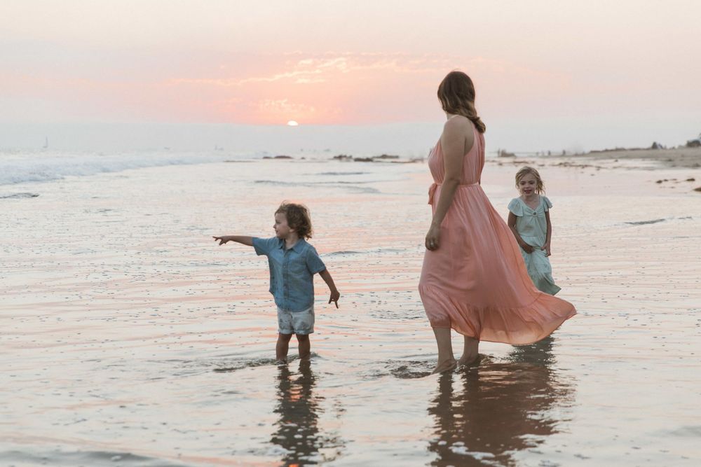 Mother in pink dress walks with two young children along shallow ocean shore at sunset.