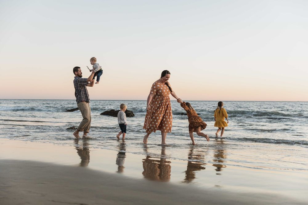 Happy family of five walking along wet sandy beach at sunset, father lifting baby.
