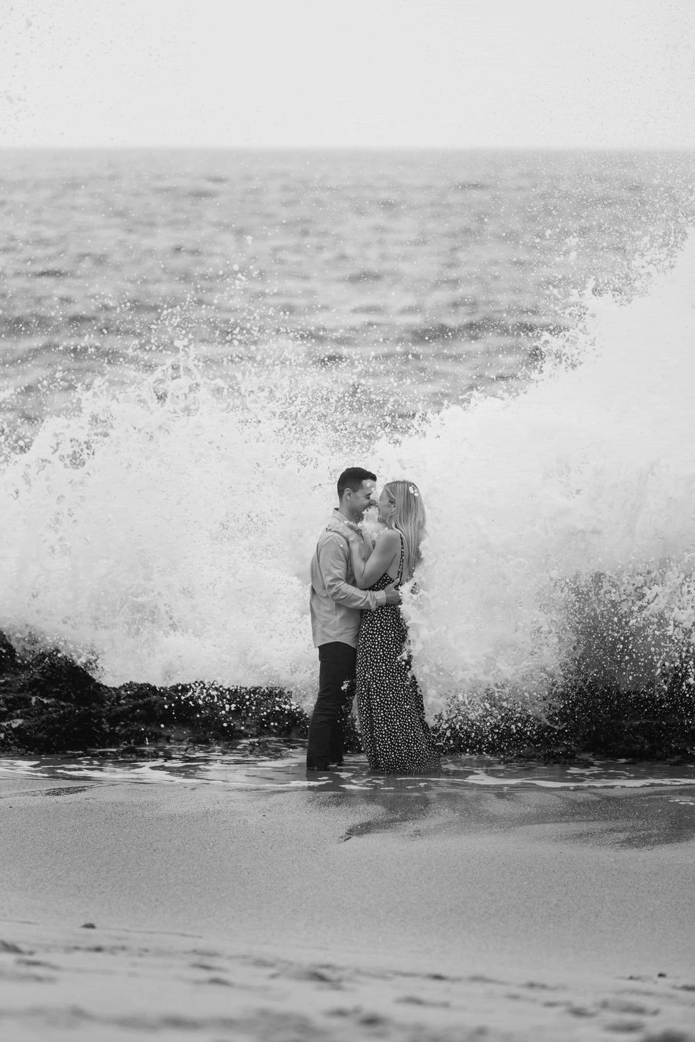 Couple embracing romantically as large ocean wave crashes dramatically behind them in black and white.