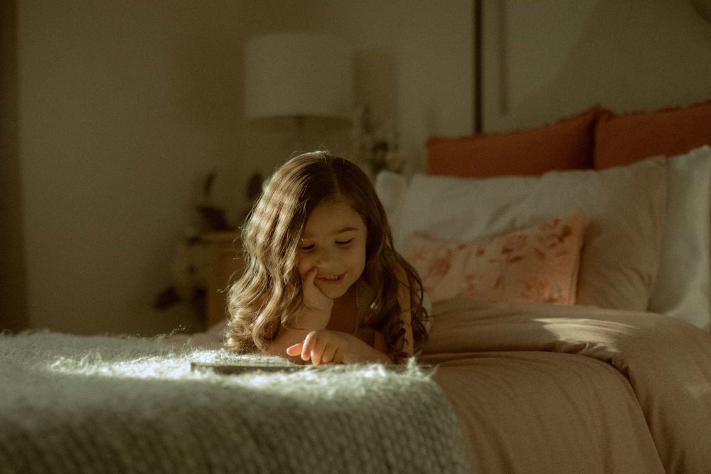 Young girl lying on bed smiling at tablet in softly lit cozy bedroom.