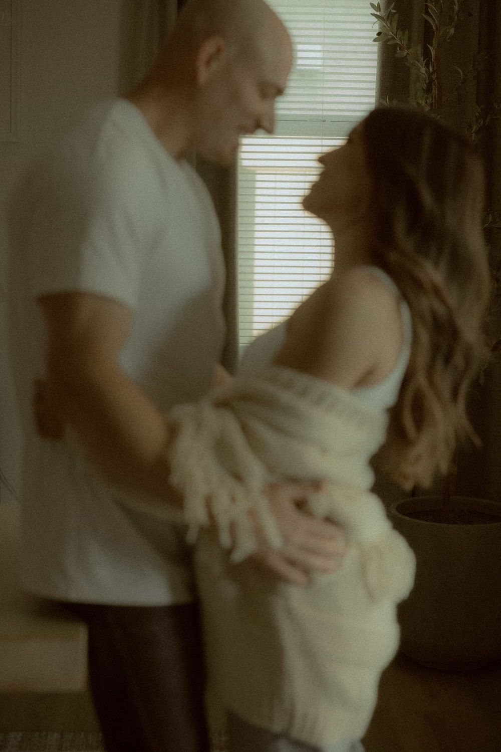 Romantic couple embracing near window with soft natural light filtering through blinds.