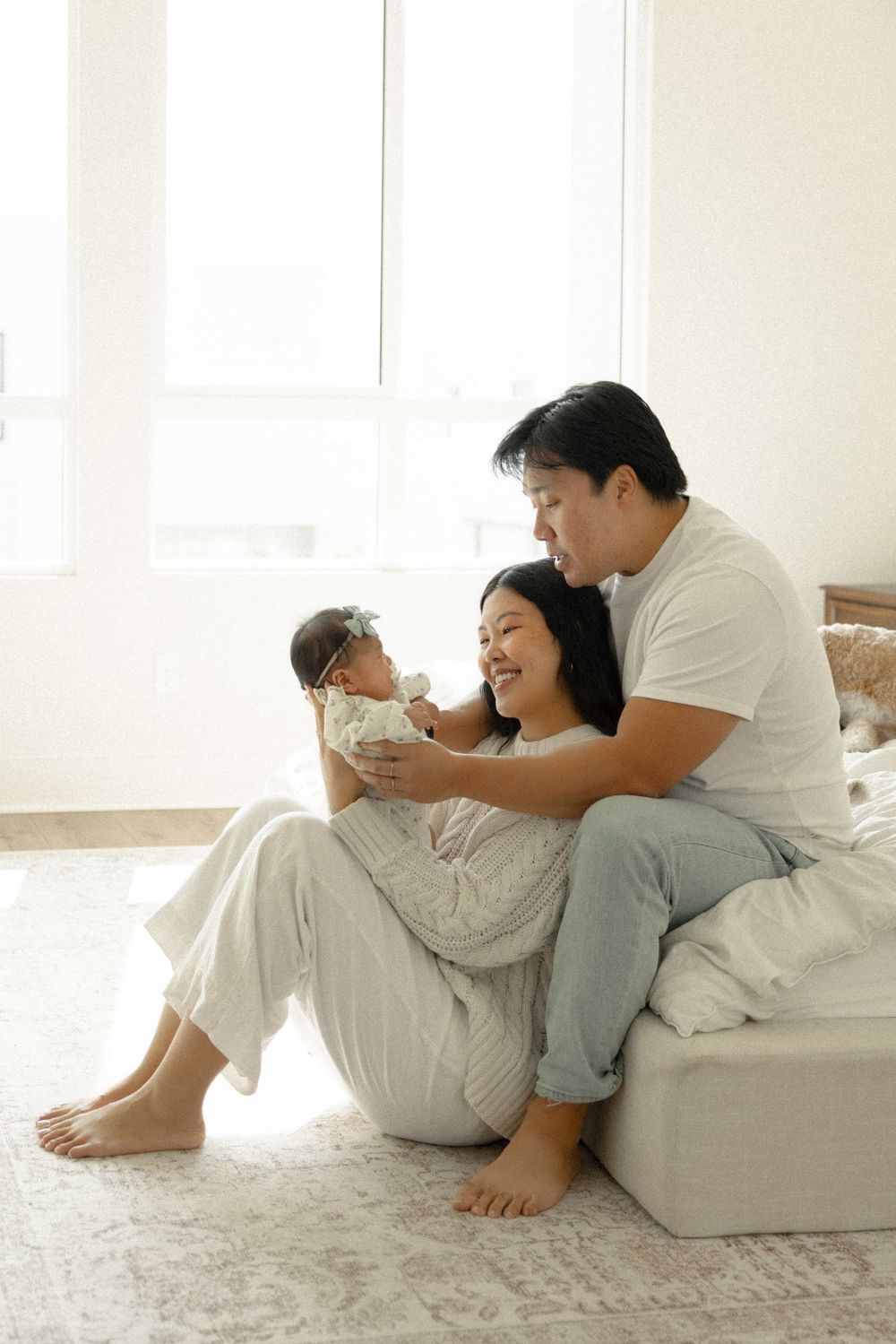 Happy Asian parents sitting on floor admiring their newborn baby in bright airy room.