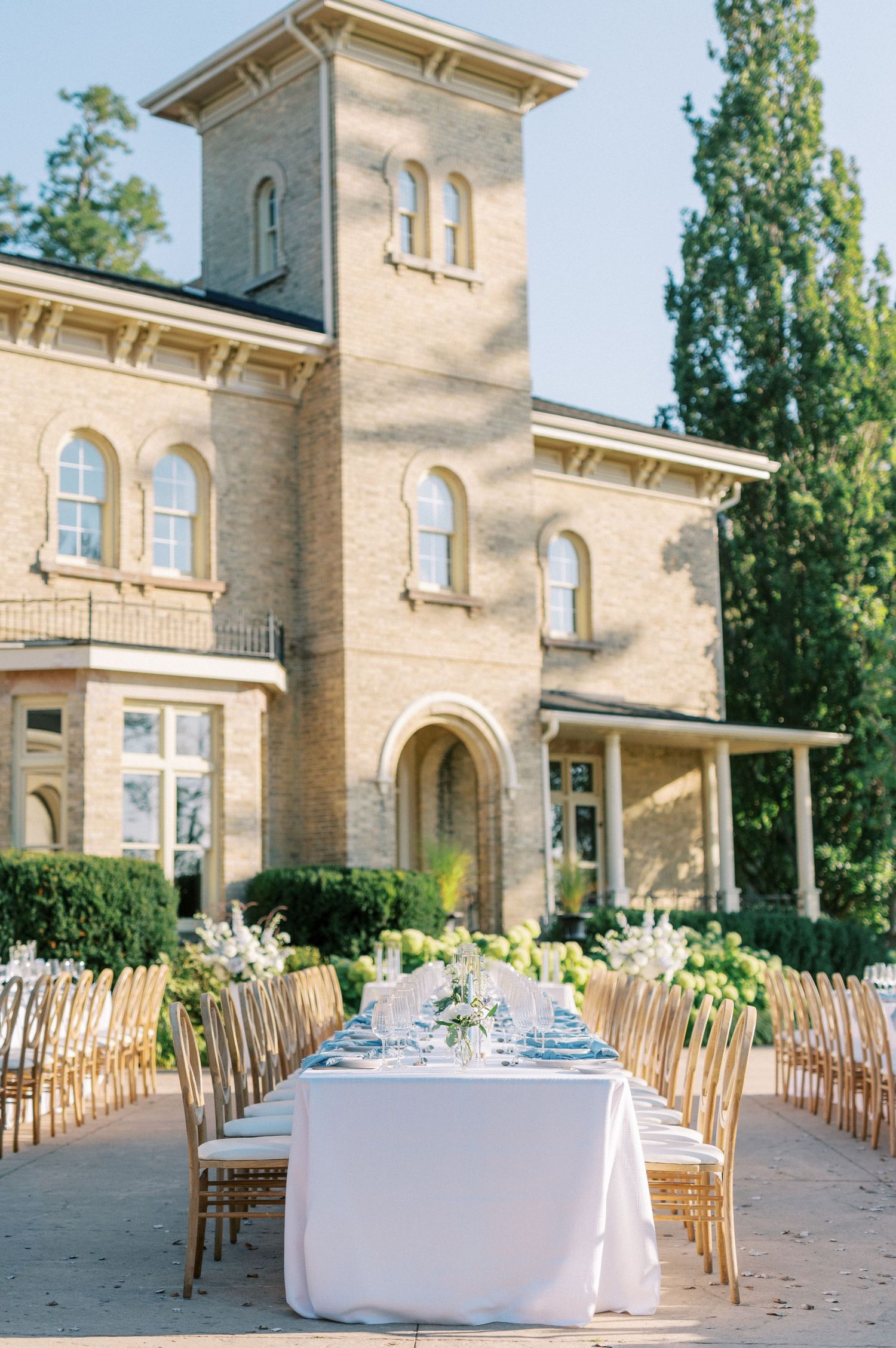 Elegant outdoor wedding reception with long white-draped tables and gold chairs set before a grand stone mansion.