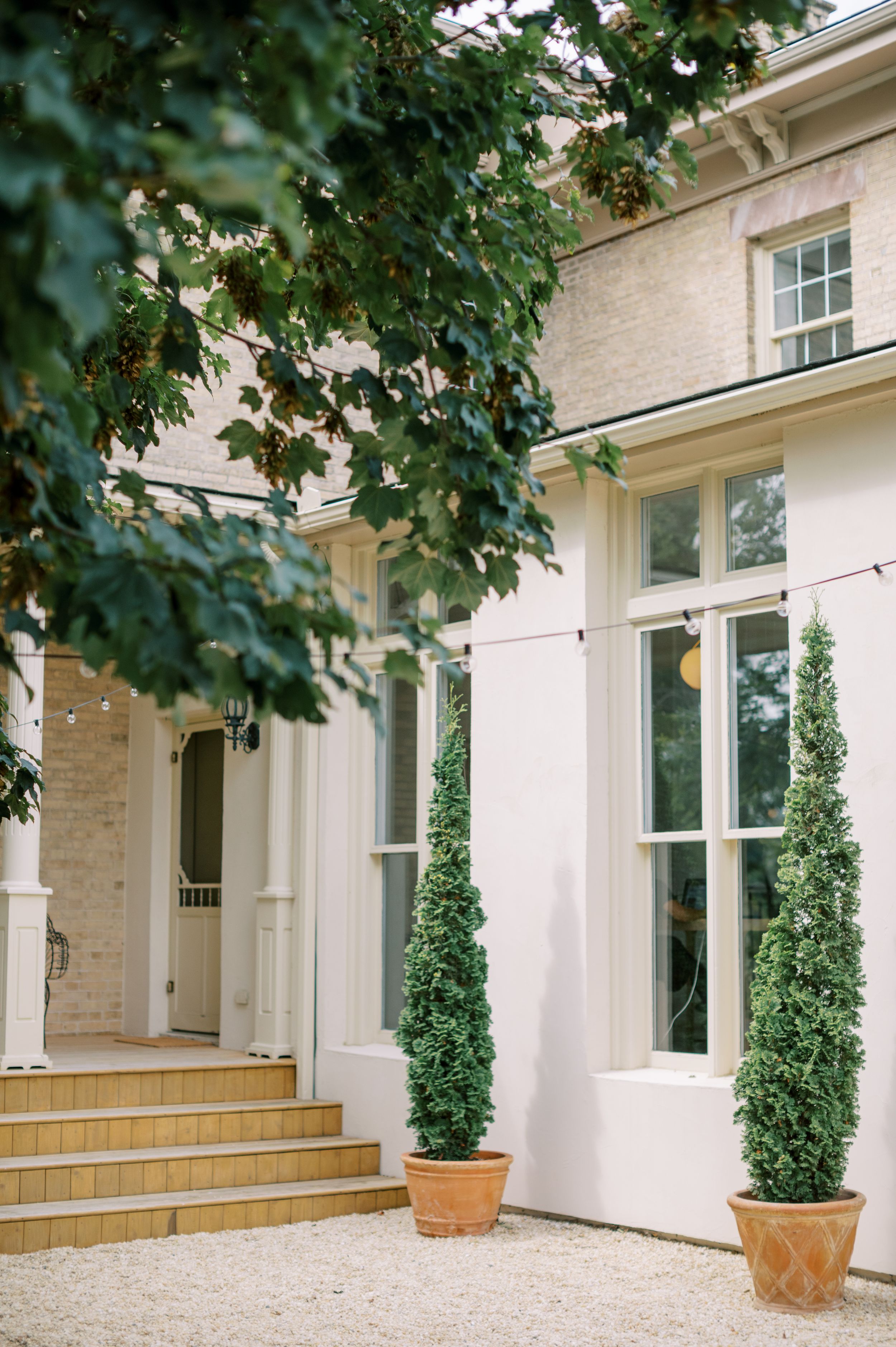 Elegant white Georgian manor house with tall topiary trees in terracotta pots, stone steps, and string lights.