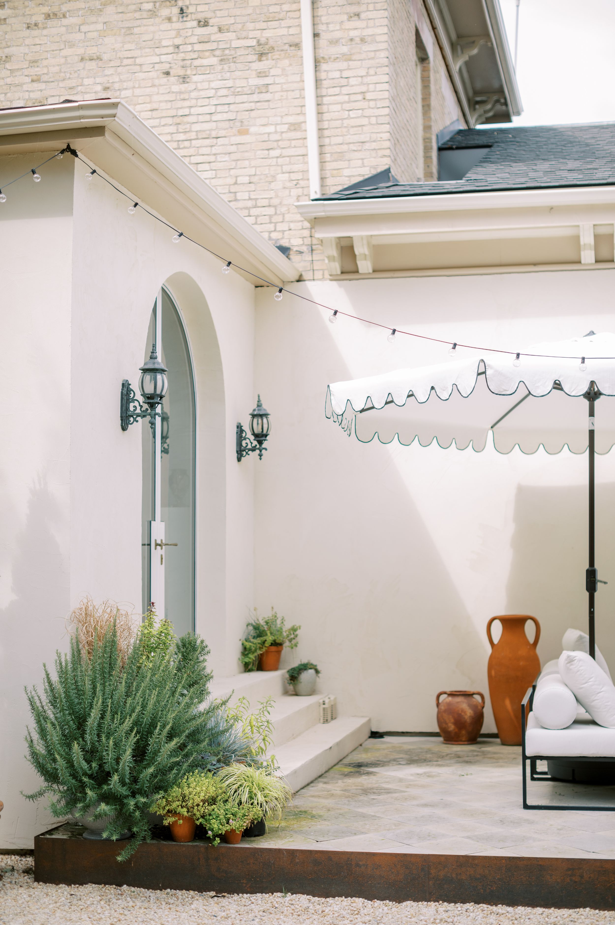 Elegant white stucco courtyard with arched door, string lights, scalloped umbrella, terracotta urns, and lush greenery.