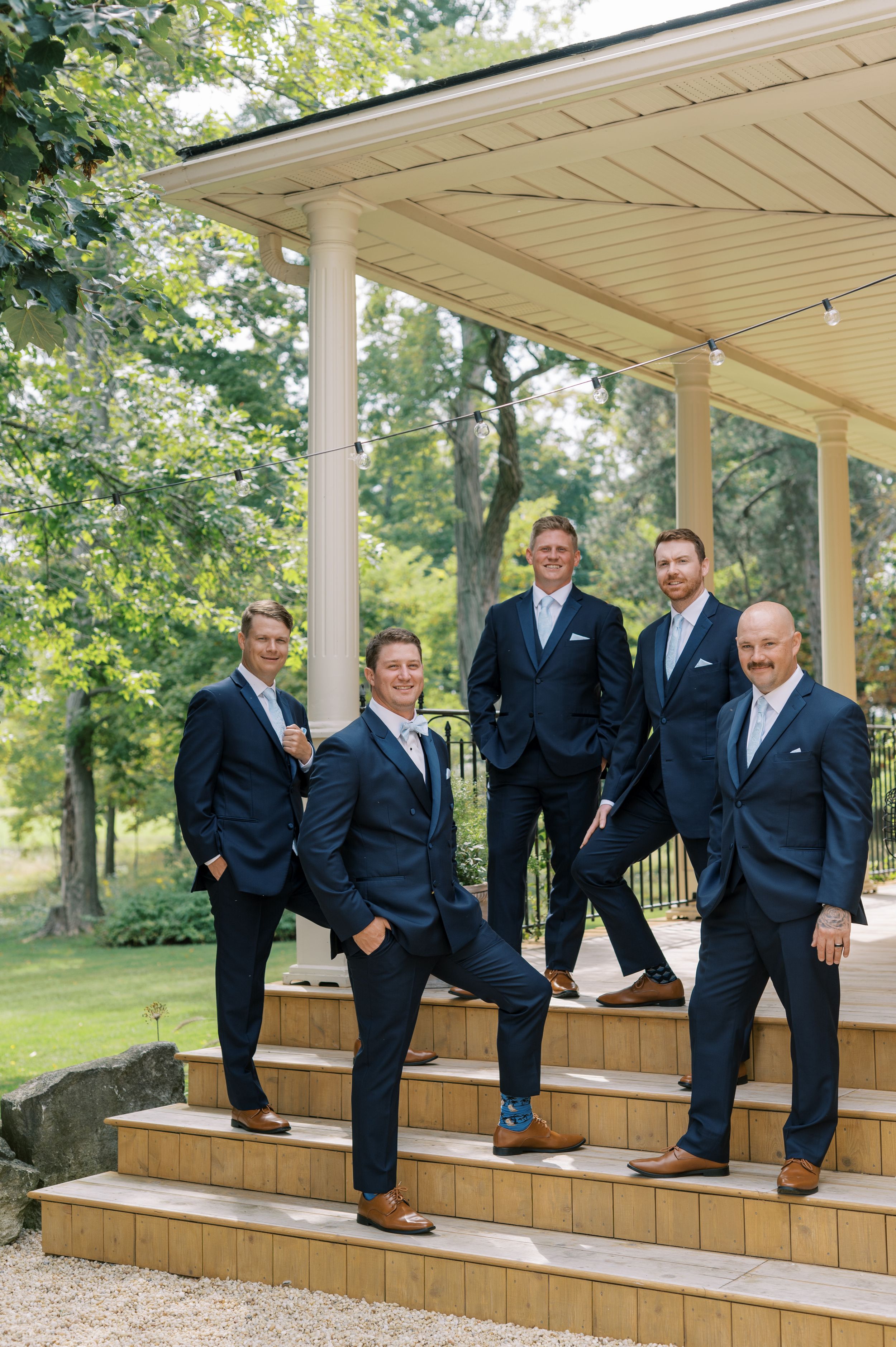 Groom and four groomsmen in matching navy suits pose confidently on outdoor venue steps for a wedding portrait.