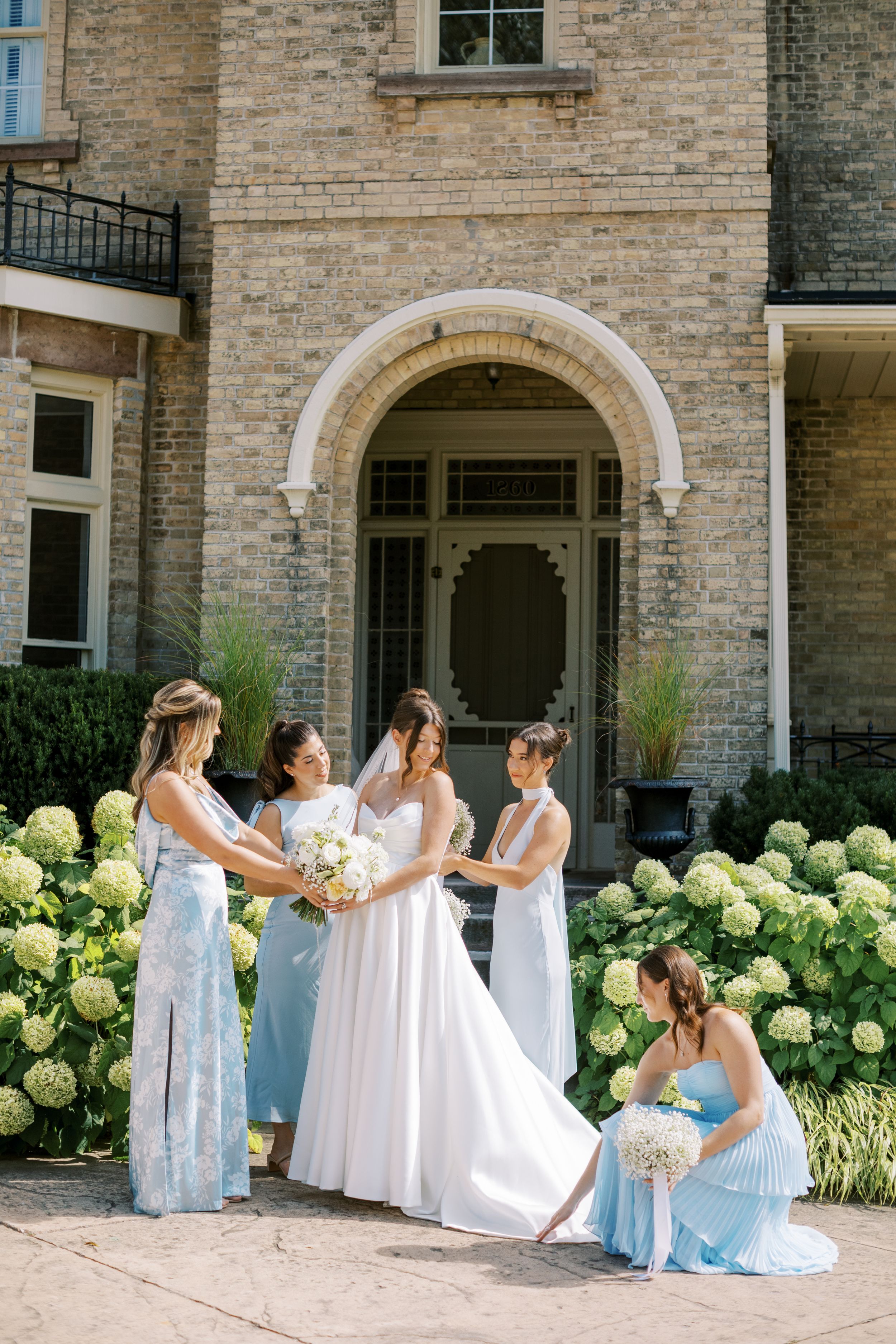 Bride in white gown with bridesmaids in light blue dresses holding bouquets outside elegant stone mansion.