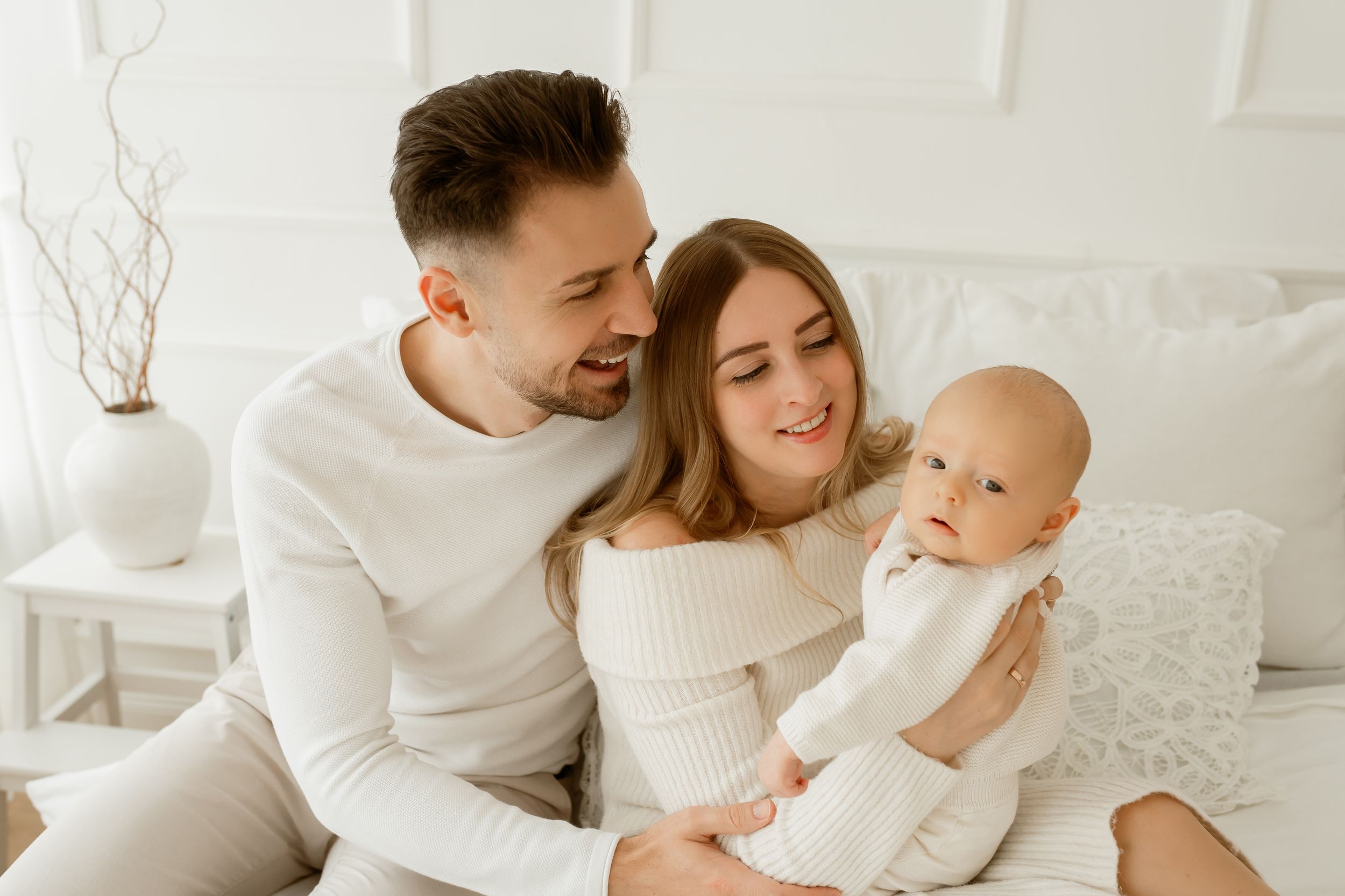 Happy young family with baby sitting on bed, parents in white outfits sharing tender moment with infant.