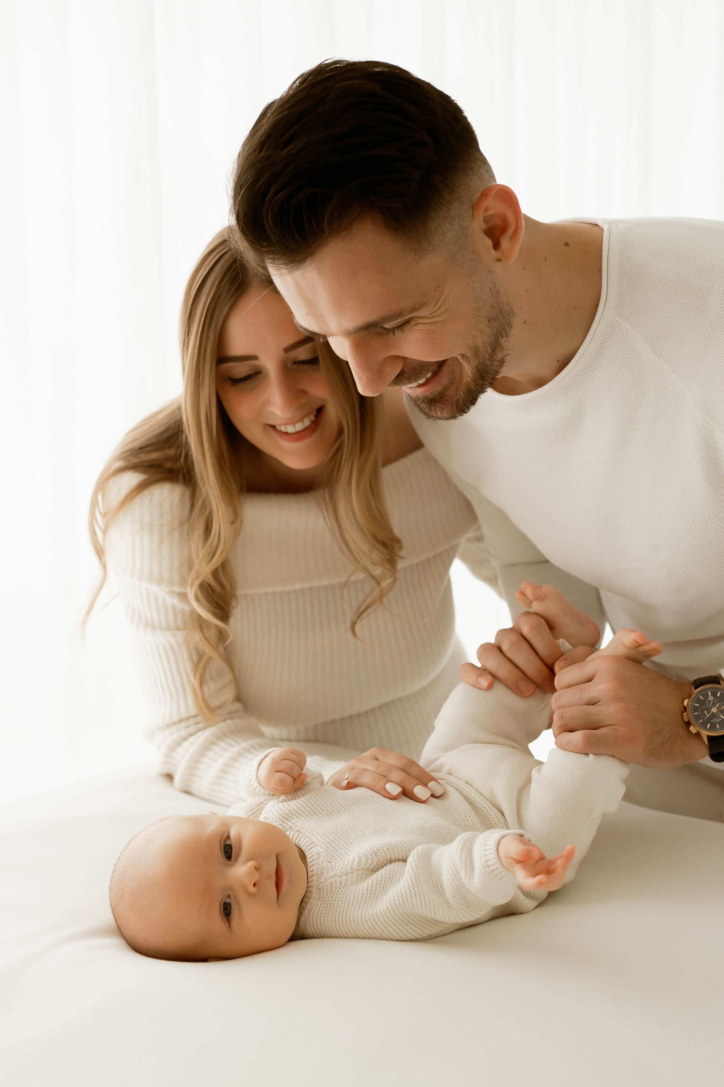 Happy young parents in white outfits smiling at their newborn baby lying on a white surface.